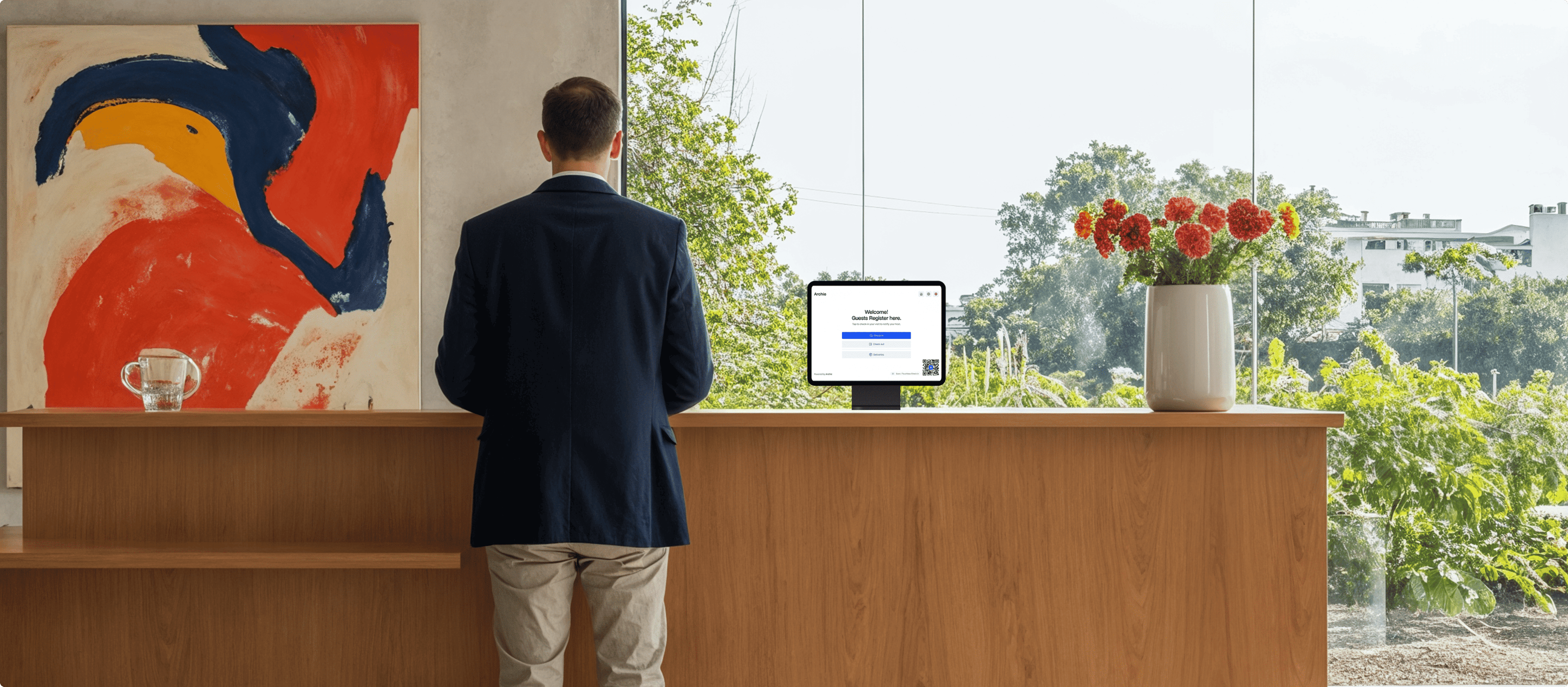 A man standing at a reception desk with a tablet displaying a guest registration screen. The desk has a vase with red flowers and a glass of water.
