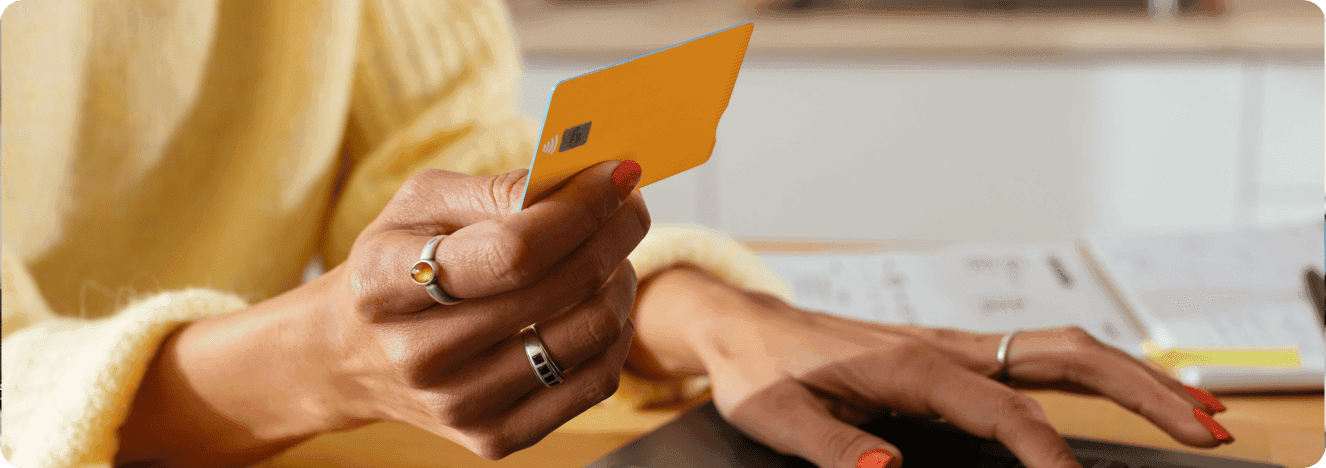 A person holding an orange payment card while using a laptop, likely making an online payment or booking.