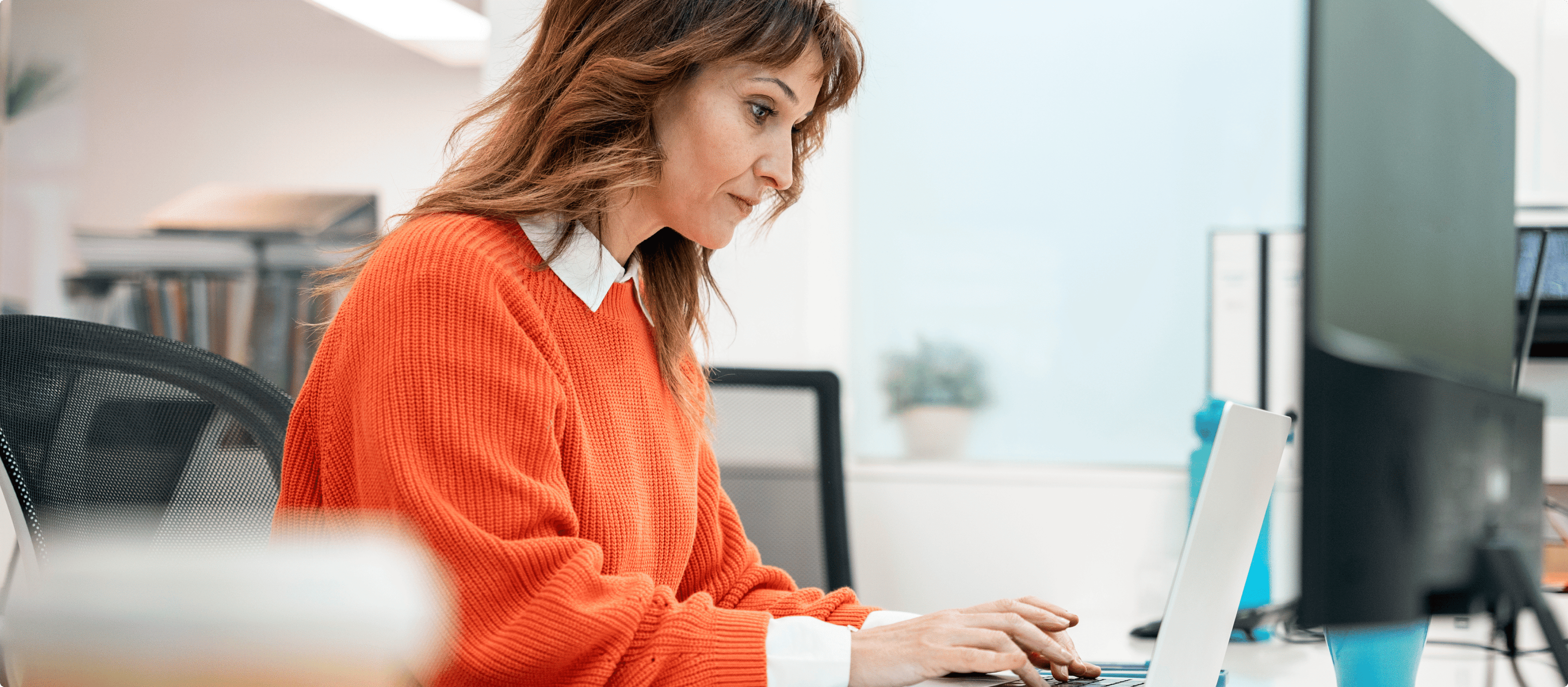 A woman with shoulder-length brown hair, wearing an orange sweater over a white collared shirt, working on a laptop in an office setting.