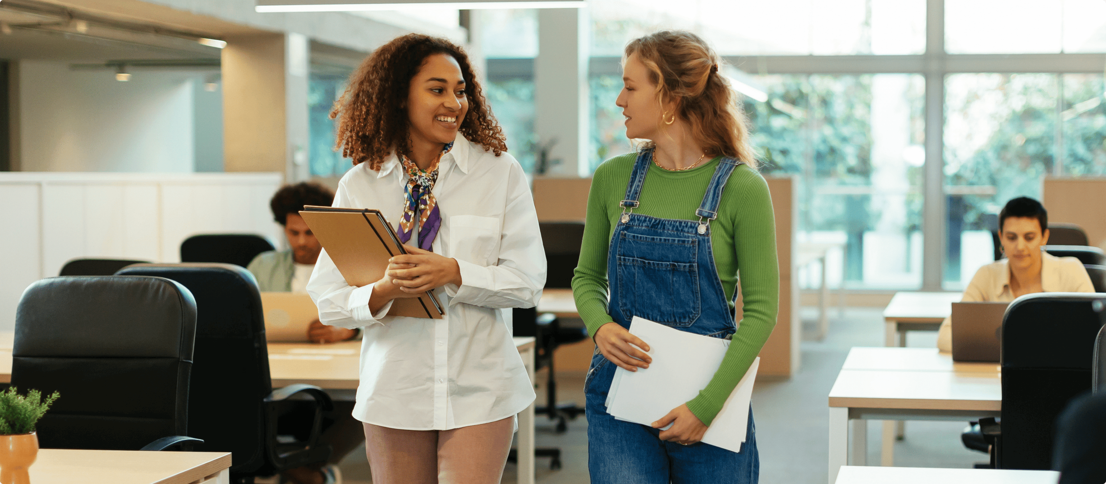 Two women in an office environment, one holding a tablet and the other holding papers, engaged in conversation. Other people are visible in the background working at their desks.