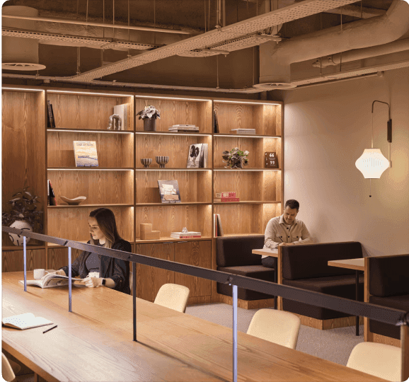 A modern co-working space with two people working at a long table, featuring wooden shelves with books and decorative items in the background.